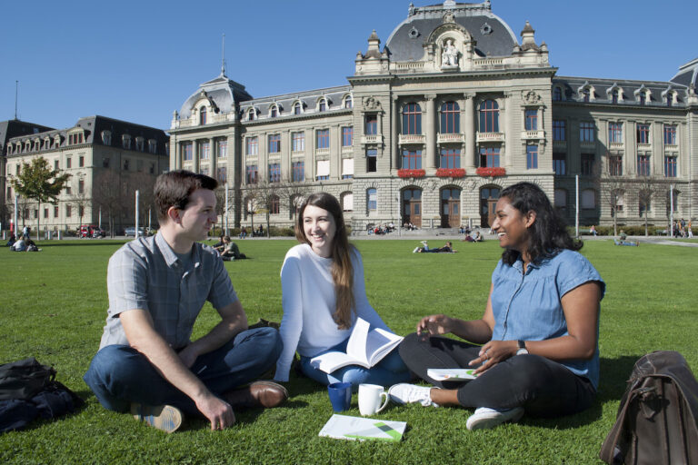 Bern Üniversitesi kampüsünde üç genç öğrenci, dinamik bir eğitim ortamında birbirleriyle etkileşimde bulunuyor.